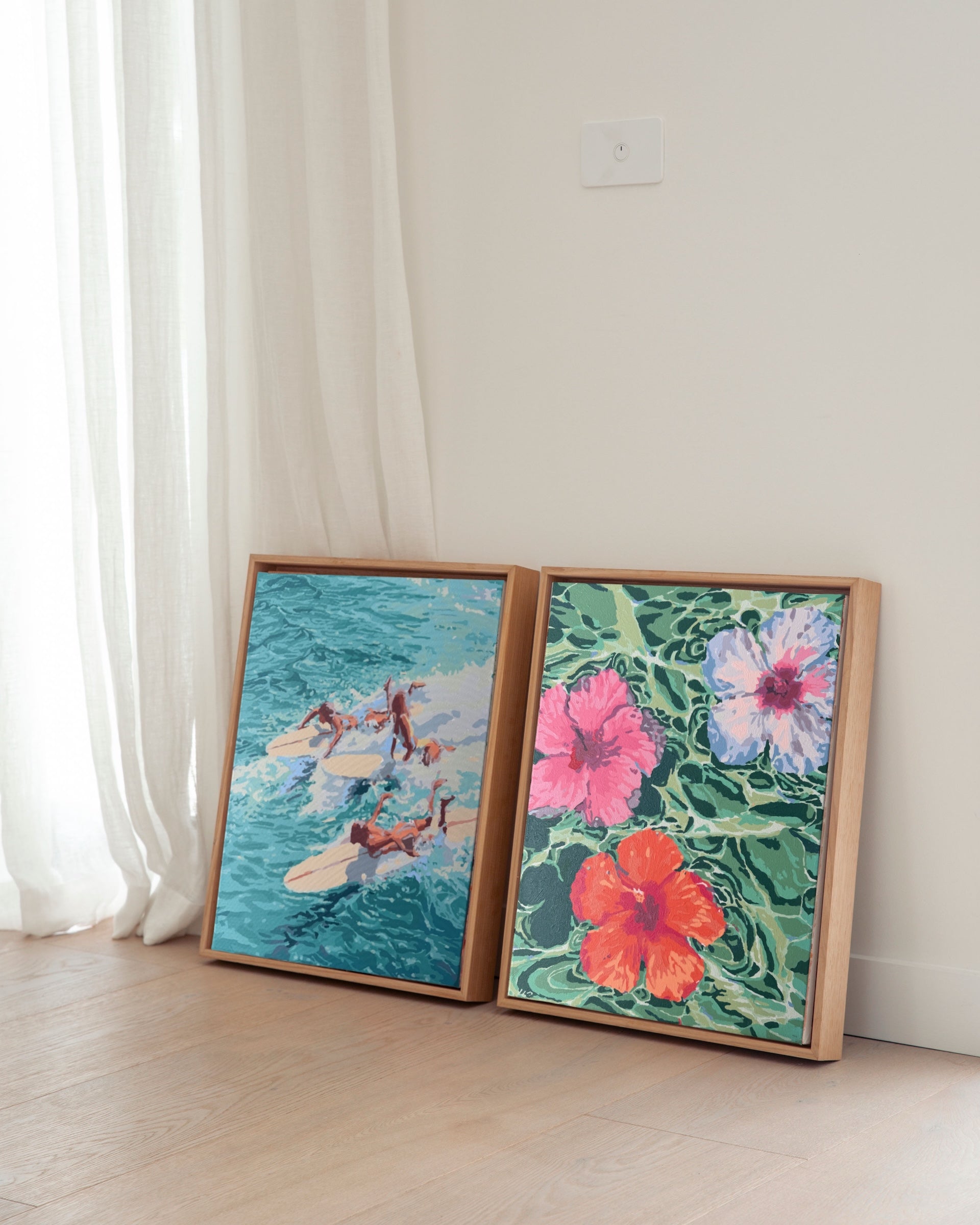 Paint Like Frida’s Hibiscus Dreams and Chaos Big Framed Canvases, displayed side by side in custom-made wooden floating frames — a bold floral scene next to an action-packed surfer moment, resting against a sunlit wall.