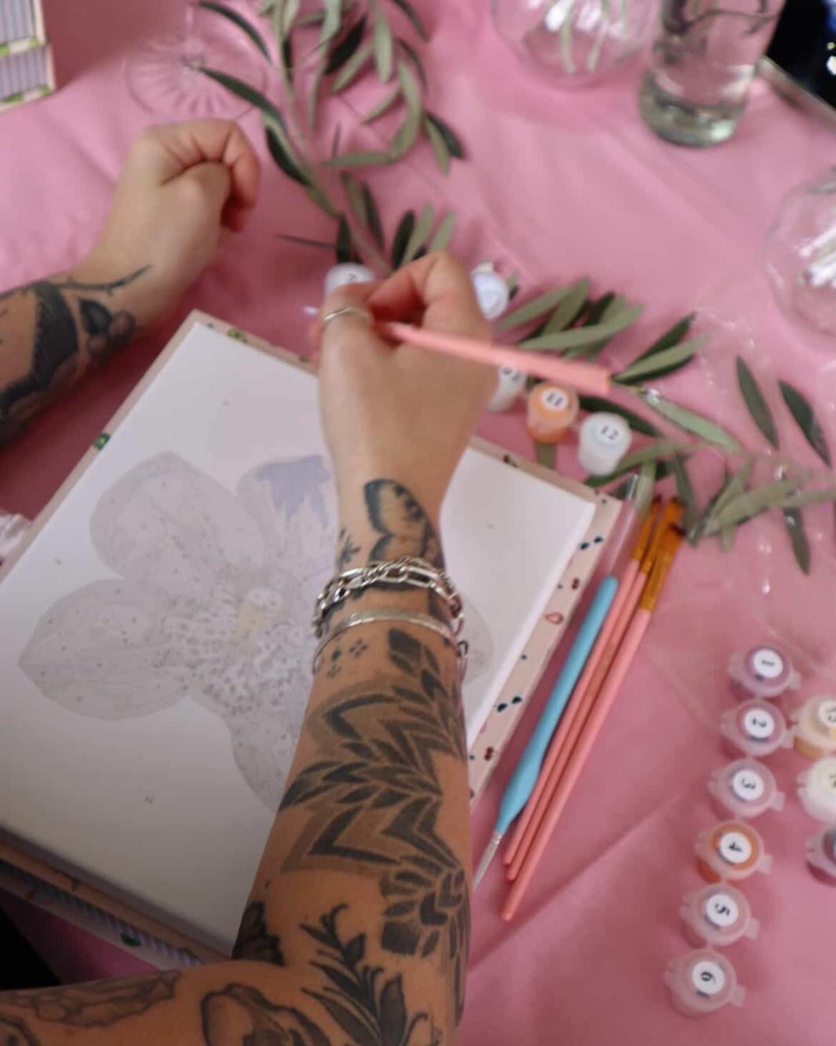 Person with tattooed arm painting an orchid flower canvas from Paint Like Frida mini framed paint-by-numbers kit at a paint and sip party, with pink tablecloth, brushes, and paint pots.
