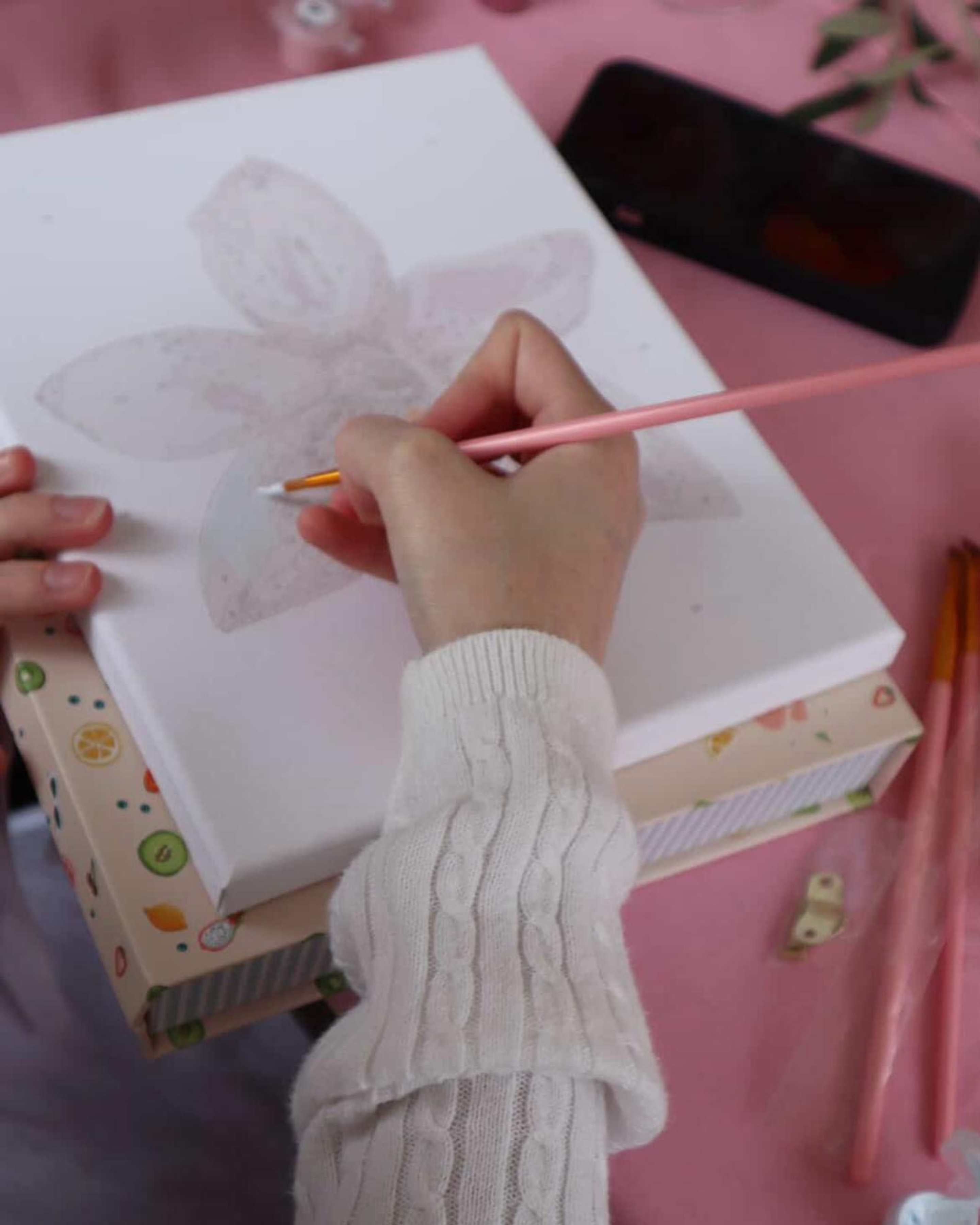 Person painting an orchid flower on a mini framed paint-by-numbers canvas from Paint Like Frida, with pink brush and pastel paints on a pink tablecloth.