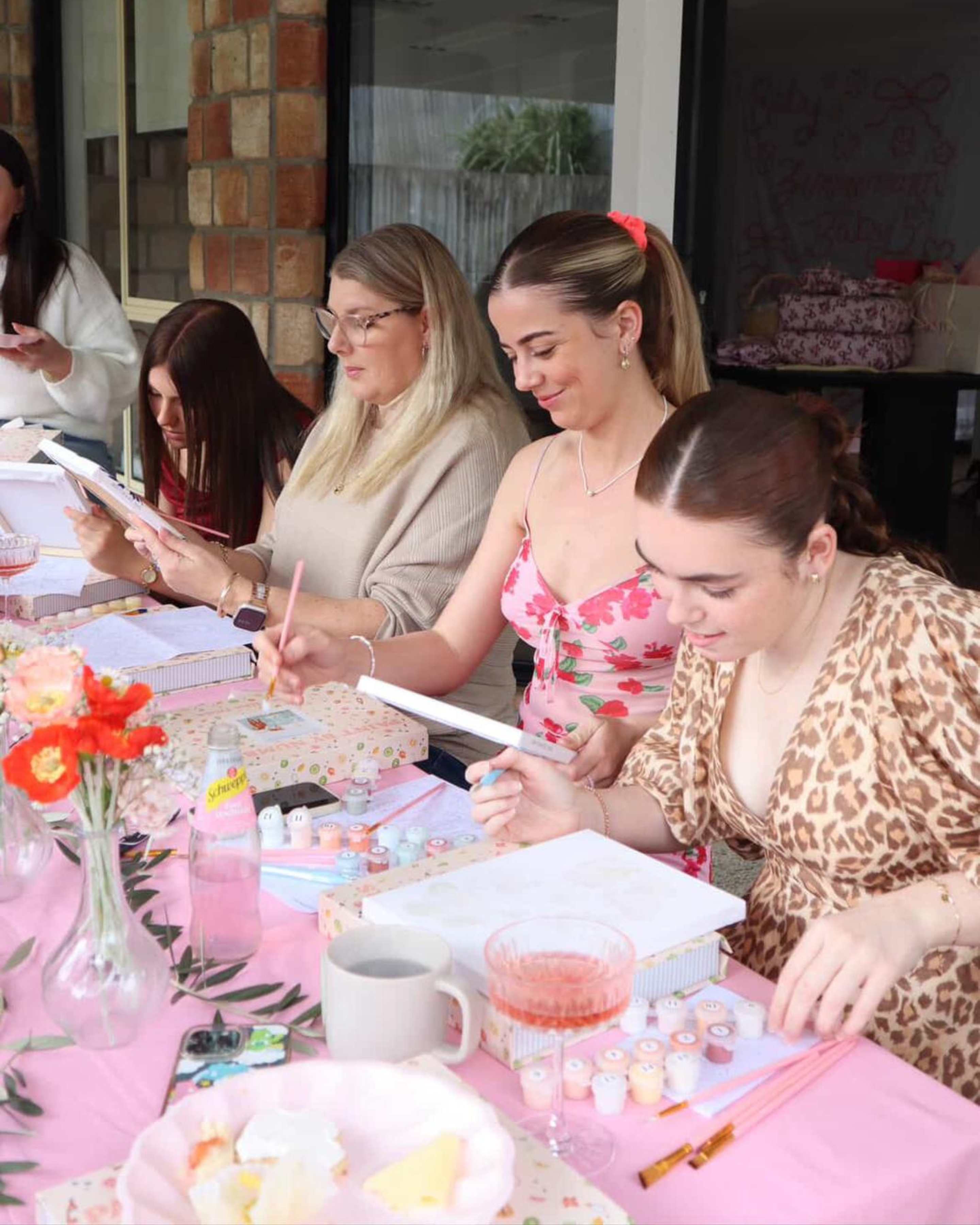 Group of women enjoying a paint and sip party with Paint Like Frida paint-by-numbers kits, pink table setting, flowers, and drinks, creating fun DIY art together.
