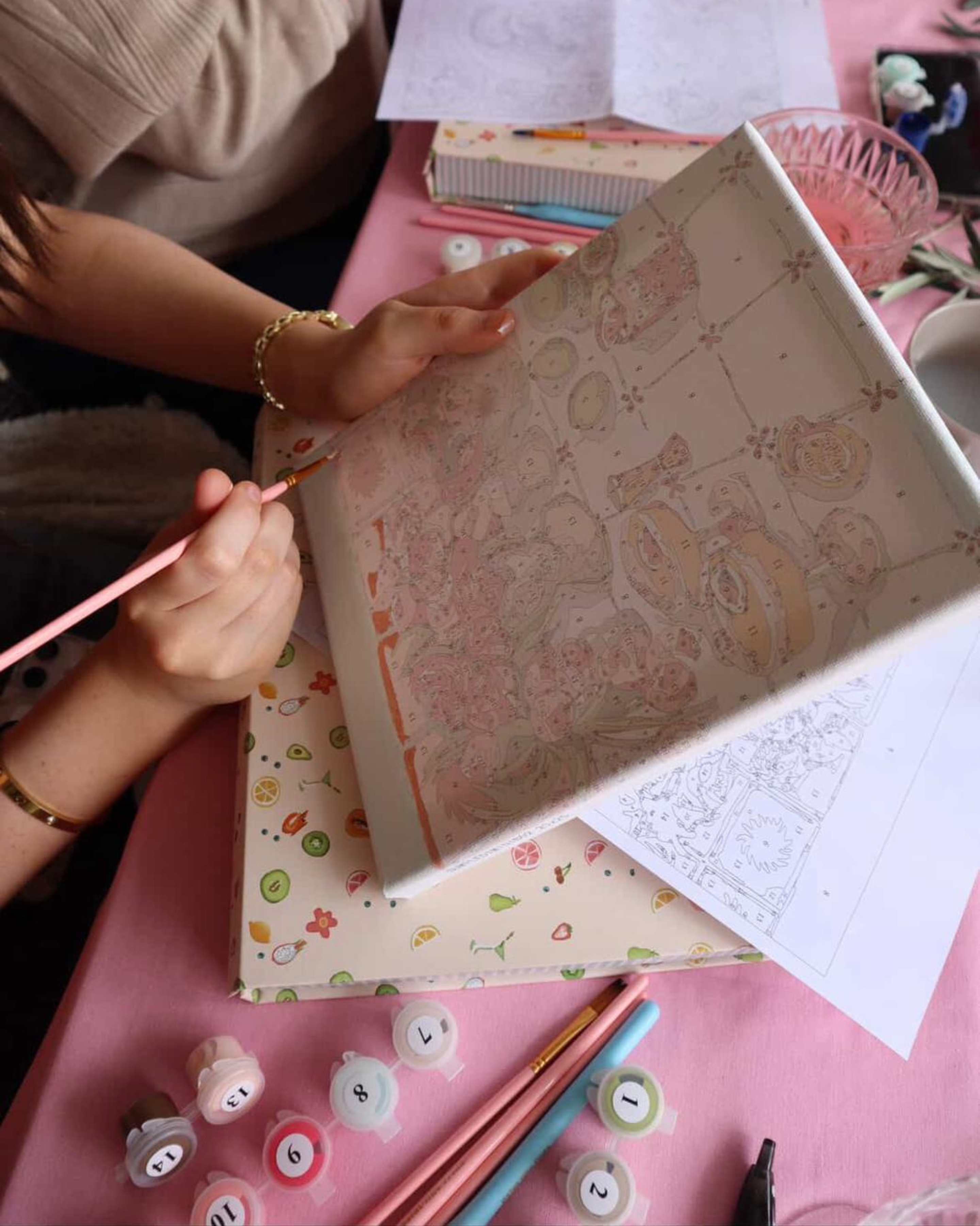 Person holding and painting a detailed Summer Fridays paint-by-numbers canvas at a fun paint and sip event, surrounded by paints and brushes.